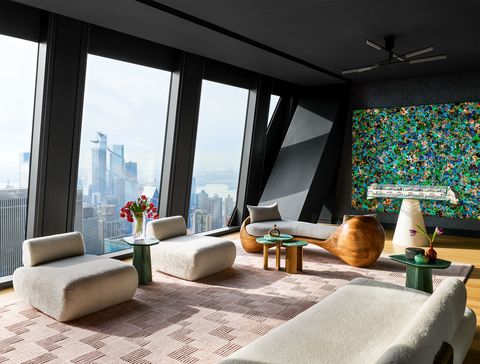 a penthouse living room has floor to ceiling windows, an armless sofa and chairs in an off white textured fabric, geometric rug, a curved wood chaise with fabric cushion, foosball table, large colorful painting