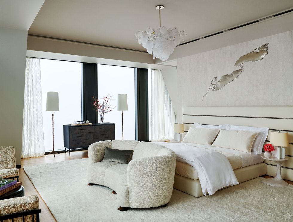 a bedroom is done in primarily off white, the slatted headboard extends beyond the bed, footed nightstands with lamps, a curved boucle sofa is at the foot, dresser flanked by floor lamps near windows