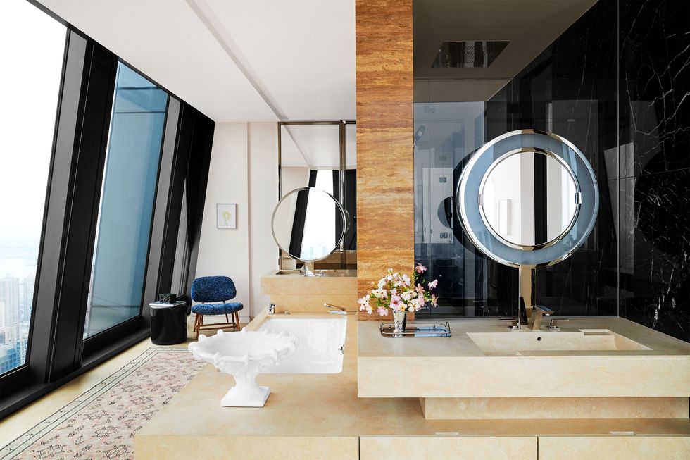 a primary bathroom has a vintage chair and side table next to floor to ceiling windows, a spa tub and two sinks built into the marble counters, two round mirrors, and a half mirrored wall