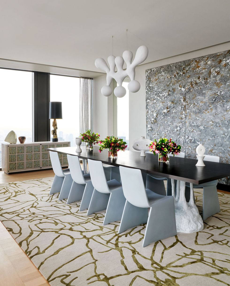 a long wood dining table with a white sculptured base, eight modern chairs in a gray molded shape, sideboard with tiles, patterned beige rug, chandelier with four shapes pointing up and glass globes below