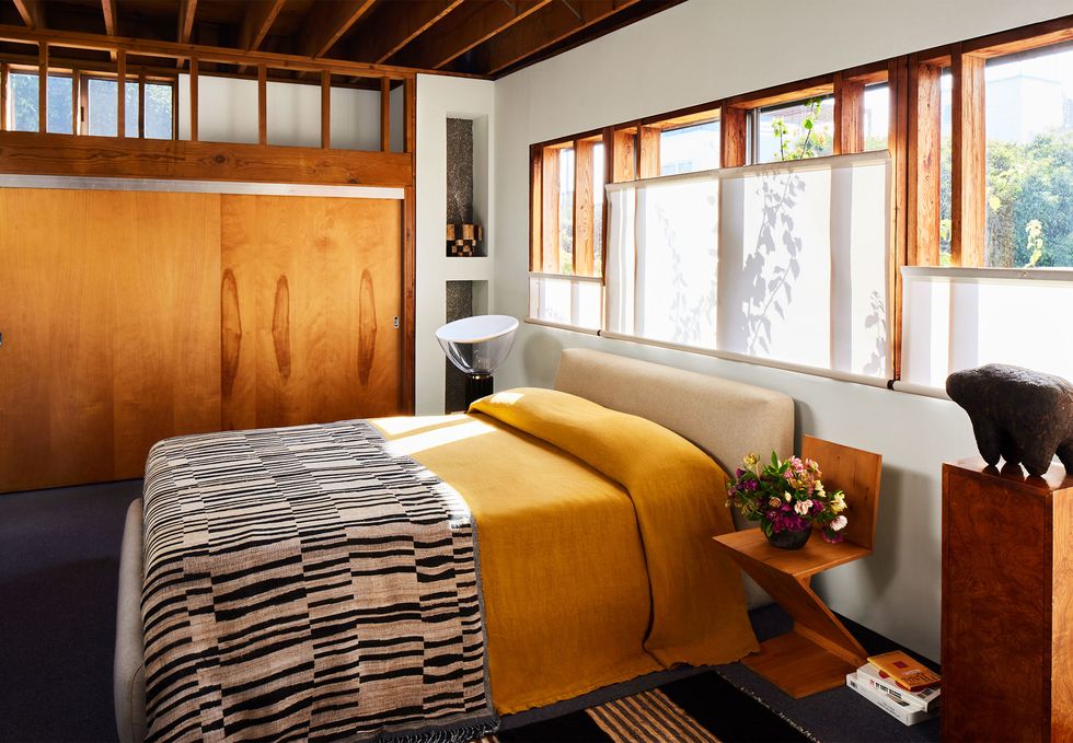 in the primary bedroom is a bed with an upholstered headboard, a bright mustard spread, and a black and white throw, a z shaped wood nightstand, windows across wall above bed, stained wood closet doors