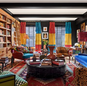 a living room has dark walls, turkish rug, bright blue sofa, club chairs with animal or geometric prints, oval cocktail tables, side tables, curtains in fuchsia, yellow, and teal, built in shelves and fireplace, large artwork above sofa