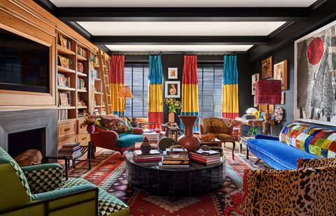 a living room has dark walls, turkish rug, bright blue sofa, club chairs with animal or geometric prints, oval cocktail tables, side tables, curtains in fuchsia, yellow, and teal, built in shelves and fireplace, large artwork above sofa