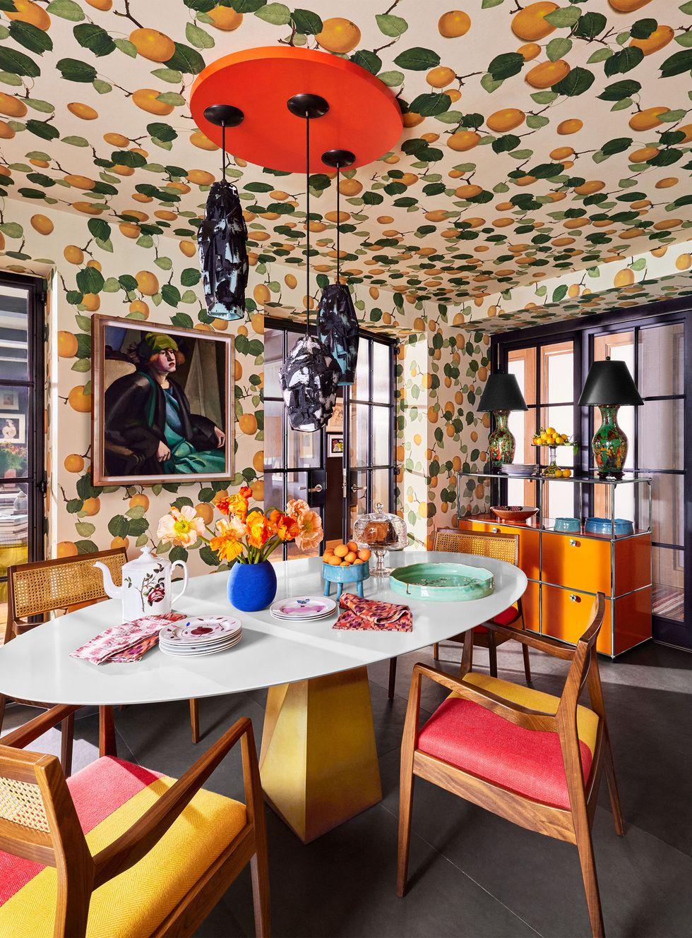 in a dining room a wallpaper in oranges and leaves print covers walls and ceiling, white oval table with four wood chairs with red and yellow cushioned seats, ceiling light with three pendants, orange sideboard with table lamps