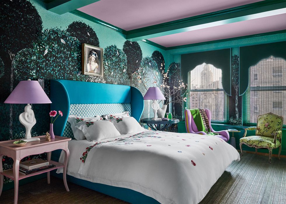 a bedroom has wallpaper with green bushes and white birds, teal fabric headboard, white bedding with small flowers, pink side table, two upholstered chairs in front of dressed windows, purple and green ceiling