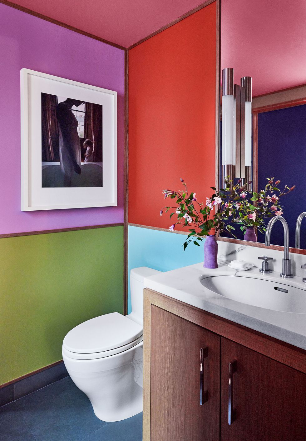 a bathroom has walls and ceiling in blocked colors of olive green, light blue, red, plum, and deep pink, a white marble vanity with wood doors, a mirror above vanity and a framed photograph on the wall