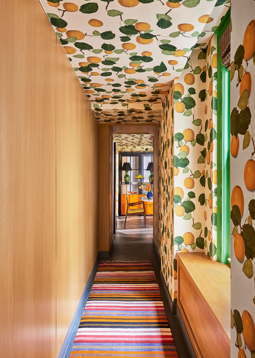 a hallway has natural wood paneling on one wall and on the ceiling and facing wall is wallpaper with leaves and citrus, a wood window seat, and striped runner