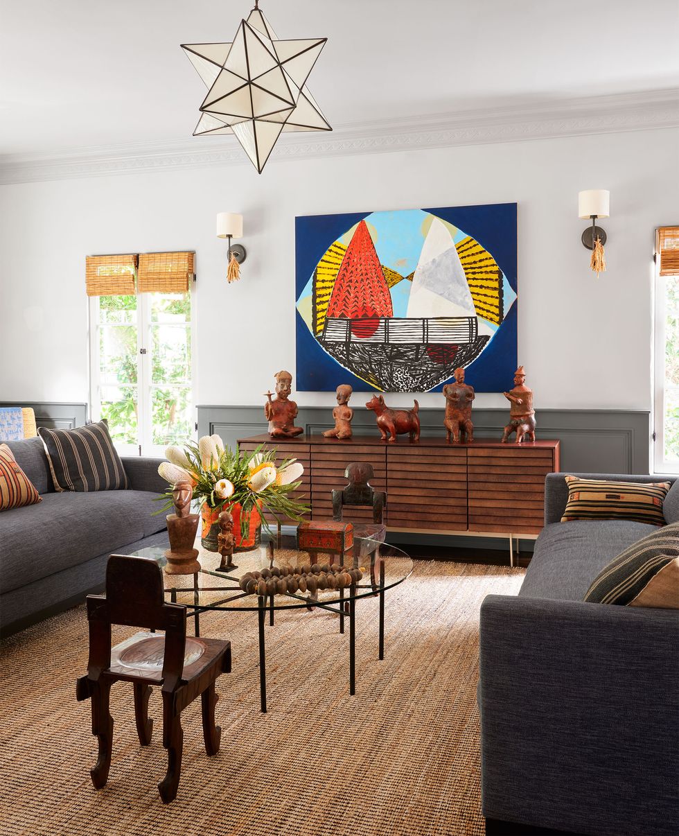 in a living room are two dark gray sofas with striped pillows facing each other, round glass topped cocktail table, a vintage wood chair, a wood sideboard with a large bright painting above, a star shaped pendant