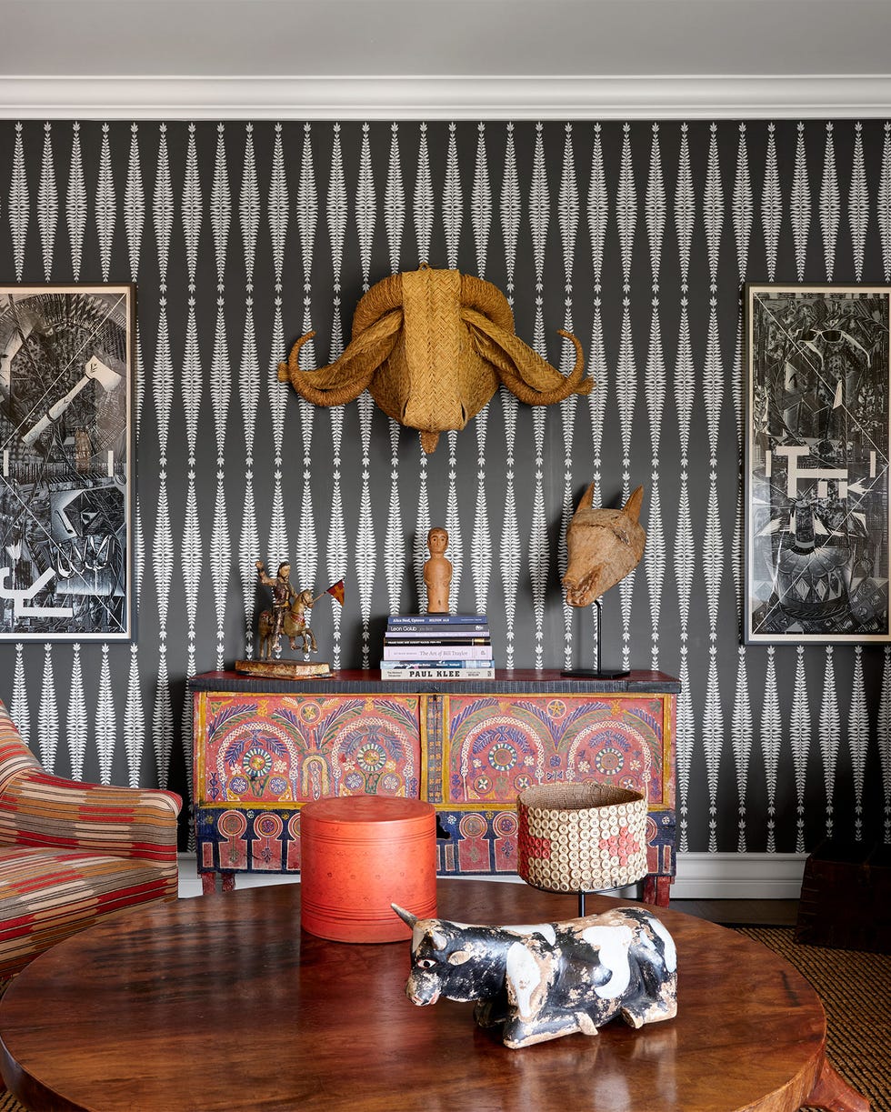 Pittman-Dowell - ELLE DECOR a sitting room has dark gray and white patterned wallpaper, a sofa, upholstered chair, cocktail table, moroccan console, a standing ceramic sculpture, and on wall a palm leaf water buffalo head and works on paper