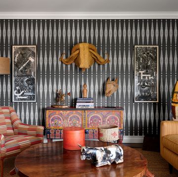 a sitting room has dark gray and white patterned wallpaper, a sofa, upholstered chair, cocktail table, moroccan console, a standing ceramic sculpture, and on wall a palm leaf water buffalo head and works on paper