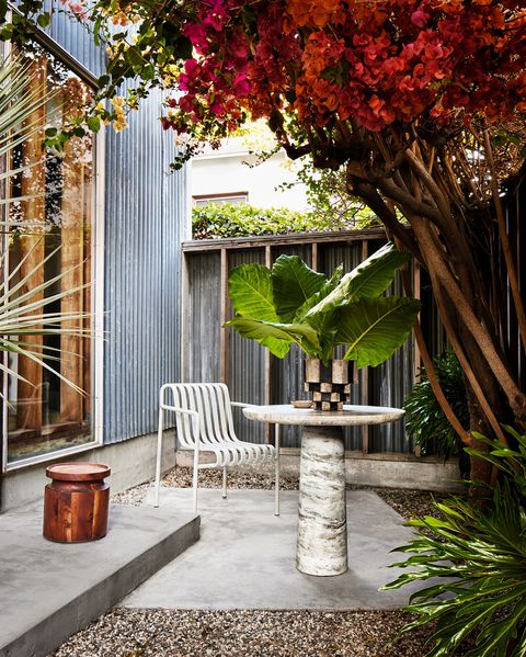 an outdoor area has a gray marble pedestal table with a planter with palm leaves, a white metal chair with curved slats, a low wooden stool, a red bougainvillea bush, a concrete floor surrounded by pebbles