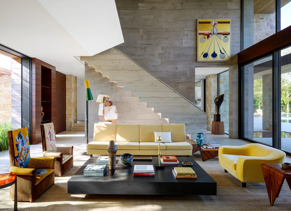 high ceilinged living room with yellow sofas and a large black low cocktail table at center with books and objets and two high back chairs on a pale yellow carpet