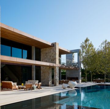 a swimming pool with a deck and lounge furniture next to a stone house with two floors with glass walls facing pool