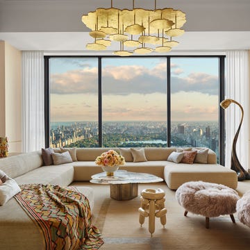 a living room with a large window overlooking central park, large sectional sofa with a colorful throw, clocktail table, chair and ottoman in fuzzy fabric, curved floor lamp, gold pendant, wall art and sculptures