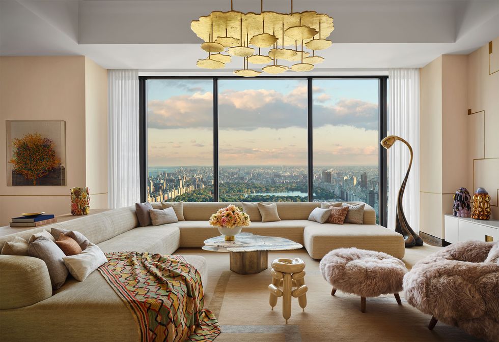 a living room with a large window overlooking central park, large sectional sofa with a colorful throw, clocktail table, chair and ottoman in fuzzy fabric, curved floor lamp, gold pendant, wall art and sculptures