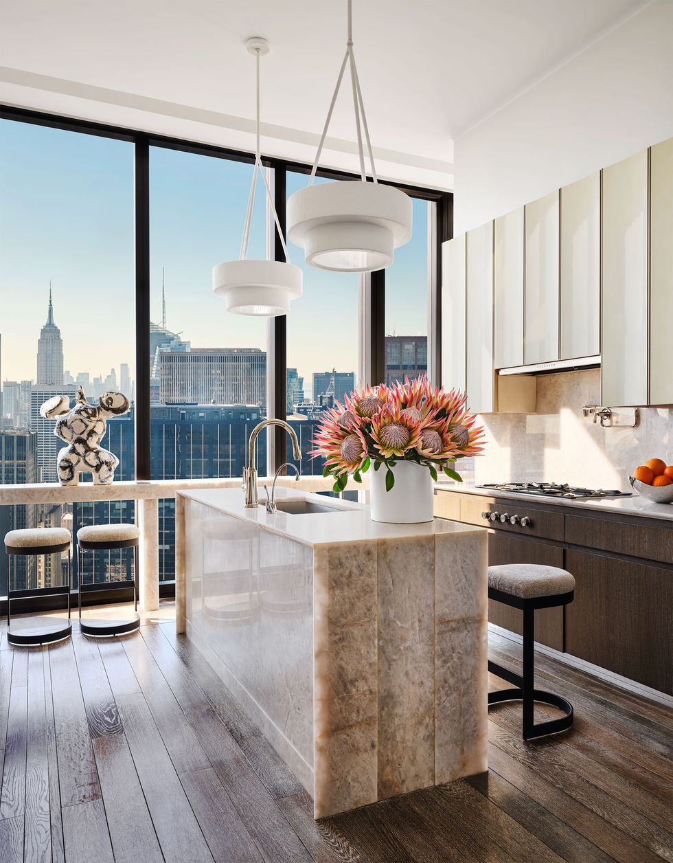 a kitchen with a large window with a narrow counter with stools and a view of skyscrapers, marble island with sink and a floral display and two pendants above, wood cabinets under a range and white cabinets above