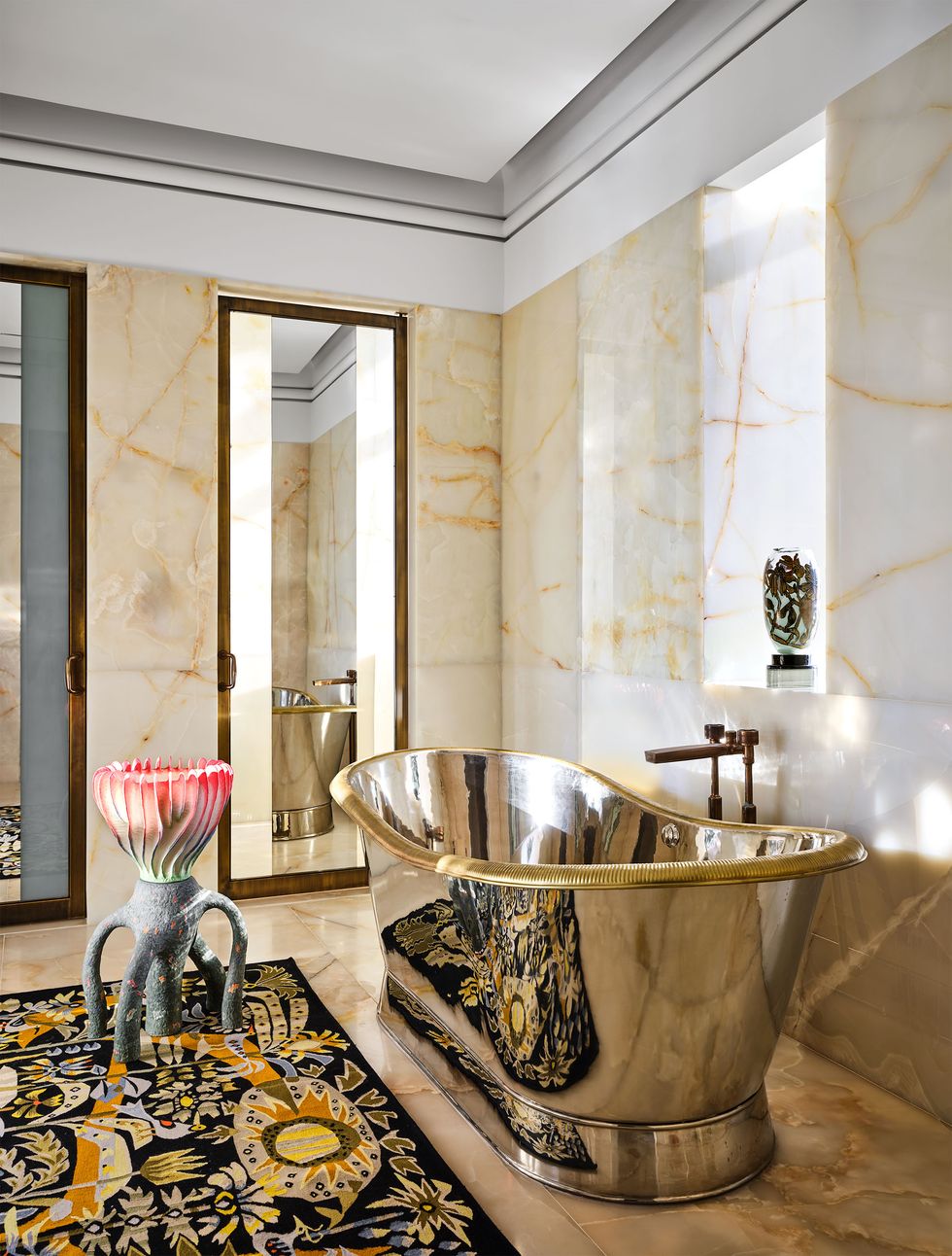 a bathroom with onyx walls with orange and yellow strands, a black background multicolored tapestry on the floor, deep, curved silver bathtub with exterior fittings, art objects on floor and window sill