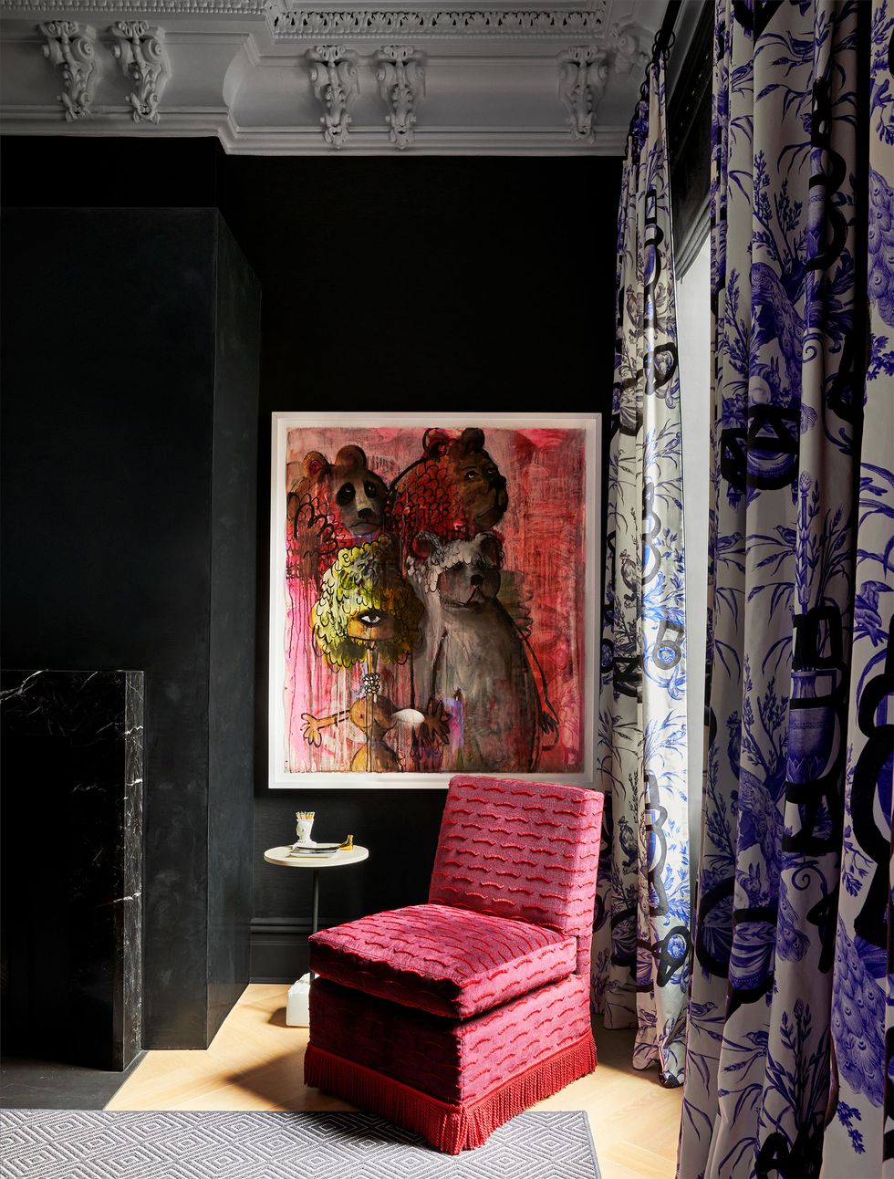 a red armless chair with fringe along the bottom in front of a painting on a wall and purple flowered long curtains