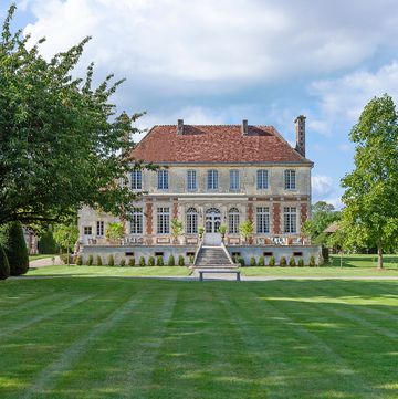 a french manor home made of limestone and brick, a lawn in front and trees along the edge, steps lead up to terraced area, two floors with large paned windows, slanted roof with chimneys