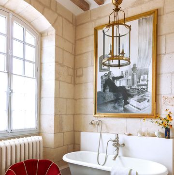 a white claw foot tub has fittings on the wall next it, glass containers and a vase on shelf behind tub and large framed artwork above, red shell chair, stool, glass pendant over tub, large window, radiator