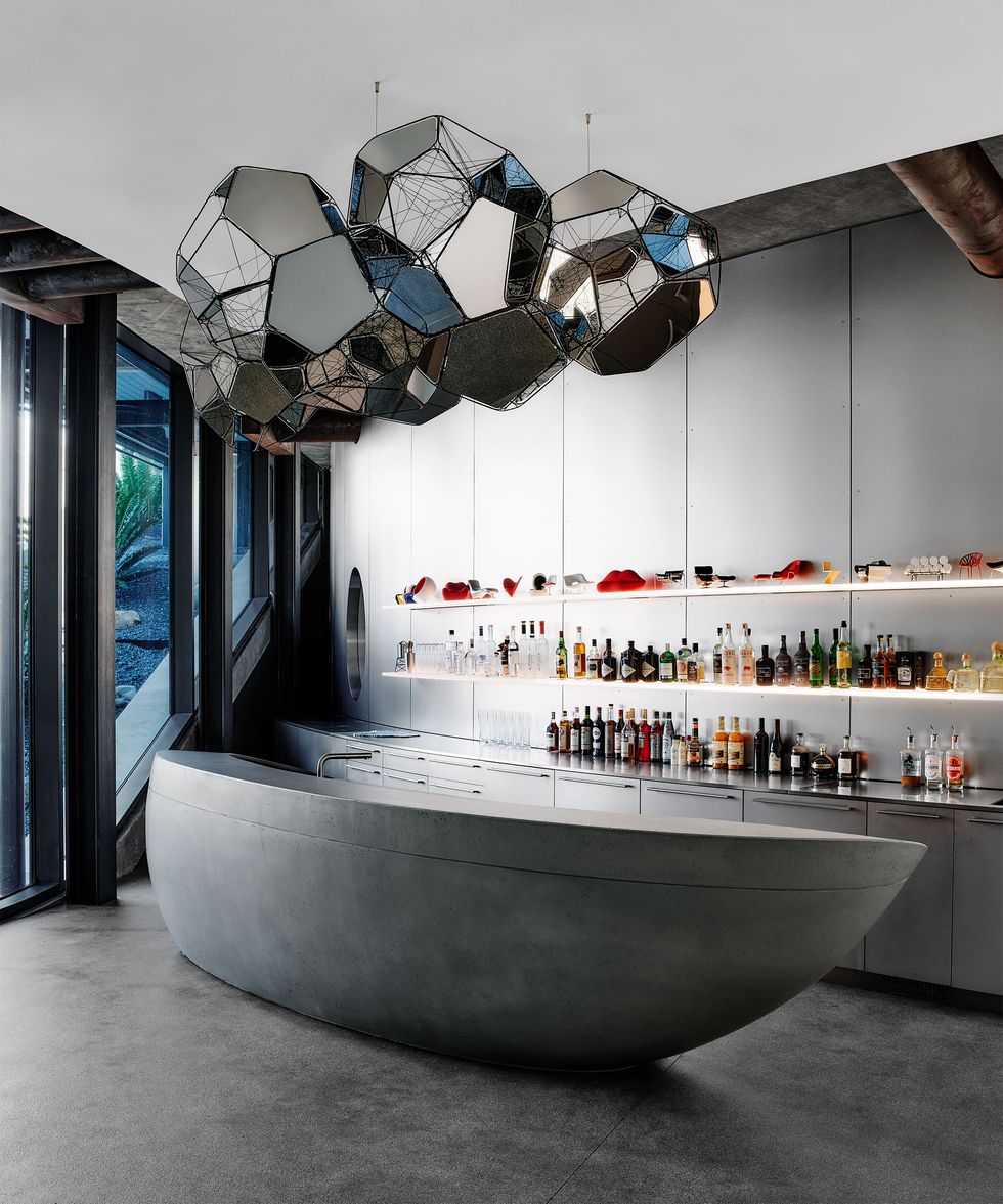 a standalone bar made of concrete shaped like a boat, concrete floor, shelves behind bar with cabinets below and bottles of spirits above, artwork with multiple reflecting facets hanging from ceiling