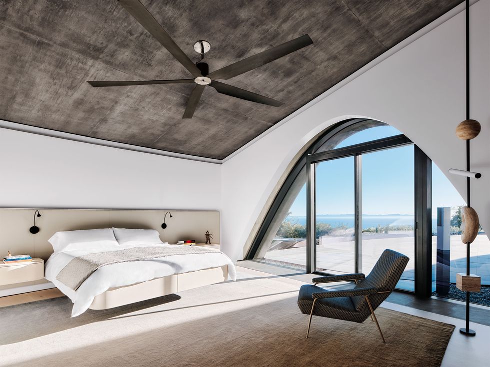 a bedroom with a high ceiling and a large ceiling fan, bed with built in extended beige fabric headboard and night stands, beige rug, armchair, a lighting sculpture, and large window with doors to terrace