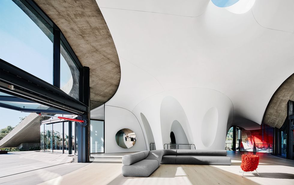 a huge room with soaring ceilings and curved white walls made of concrete, arched doorways, curved wall of glass, large gray right angled sofa, wrapped orange chair, wood floor