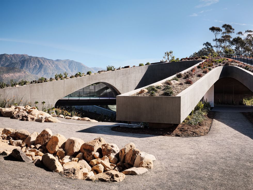 the exterior of the house with concrete rectangular wings emanating from an elevated center, green roof gardens atop each wing, blue sky and mountains in the distance, and a driveway lined with rocks