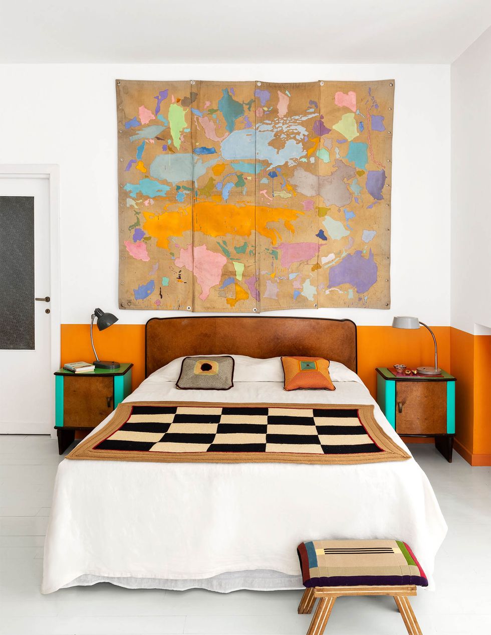 a white walled bedroom with bottom third painted orange, wood headboard and nightstands with aqua sides, gooseneck lamps, white bedcover with checkerboard throw, footstool, artwork in pastels