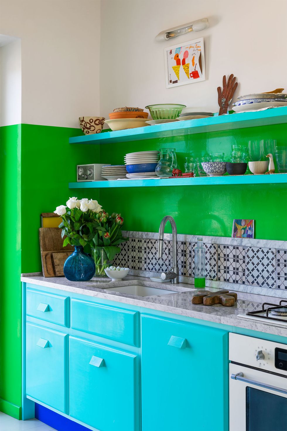 a kitchen with half white and half bright green walls, bright aqua cabinets with a deep blue toe kick, oven, marble counter with sink and range, tiled backsplash open green shelves with dishes, small artwork above