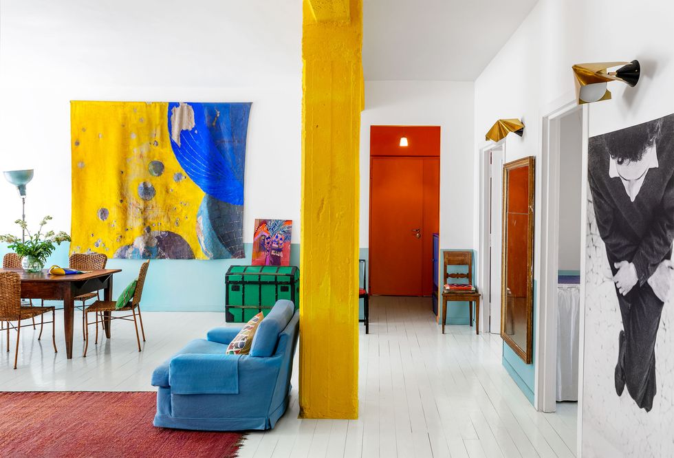 a blue sofa, bright yellow column, white and light blue walls, white wood floor, deep red rug, red entry door, green trunk, bright yellow and blue fabric painting, dining table and chairs, two sconces, other artworks