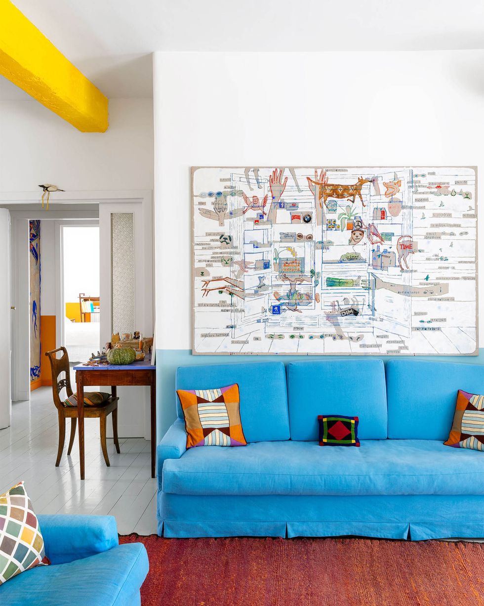 half white and half light blue walls, white wood floor covered with a deep red rug, blue sofa and matching chair both with decorative pillows, small desk and chair, large artwork over sofa, yellow ceiling beamjulie polidoro elle decor