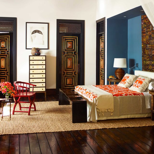 bedroom with dark floors and intricate carved and painted doors and a inset bed on a sisal rug and a red chair and slim highboy in the back