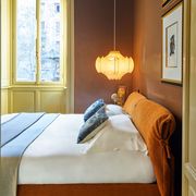 primary bedroom door opens to a bed with an orange hued fabric headboard and base with white linens and colored throw pillows, a pendant hangs over it, walls are brown and window trim is green beige