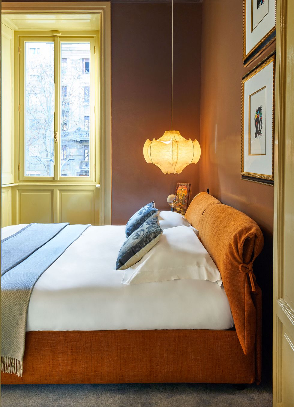 The door to the master bedroom opens to reveal a bed with an orange fabric headboard and a base with white linens and colored cushions, a pendant hanging above it, brown walls, and a window. The trim is green beige