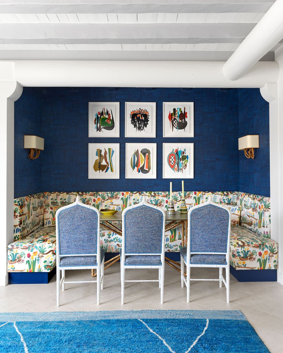 dining table set into an alcove where fabric cushioned chairs face a three sided banquette covered in a spring print fabric, two sconces and colorful framed artwork hang on deep blue walls behind the banquette