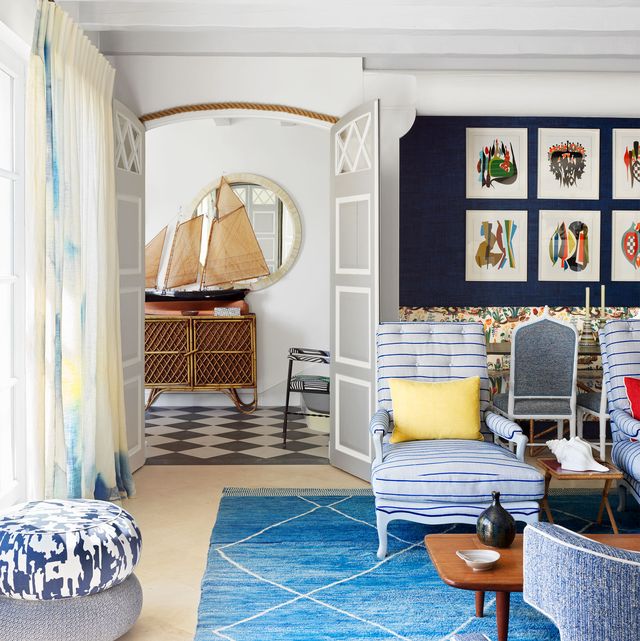 living room with a pouf in blue printed fabric next to tall curtained windows, a blue area rug, two blue and white striped armchairs, cocktail table, and a sofa, doors lead to a foyer where a model sailboat sits atop a console