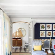 Jean-Louis Deniot - ELLE DECOR living room with a pouf in blue printed fabric next to tall curtained windows, a blue area rug, two blue and white striped armchairs, cocktail table, and a sofa, doors lead to a foyer where a model sailboat sits atop a console