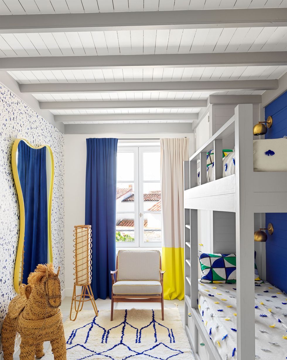 a kids’ bedroom has bunkbeds with drawers and white spreads with yellow and blue pompoms, blue and white wallpaper and area rug, a chair and floor lamp in front of a curtained window, a large mirror and a rope donkey