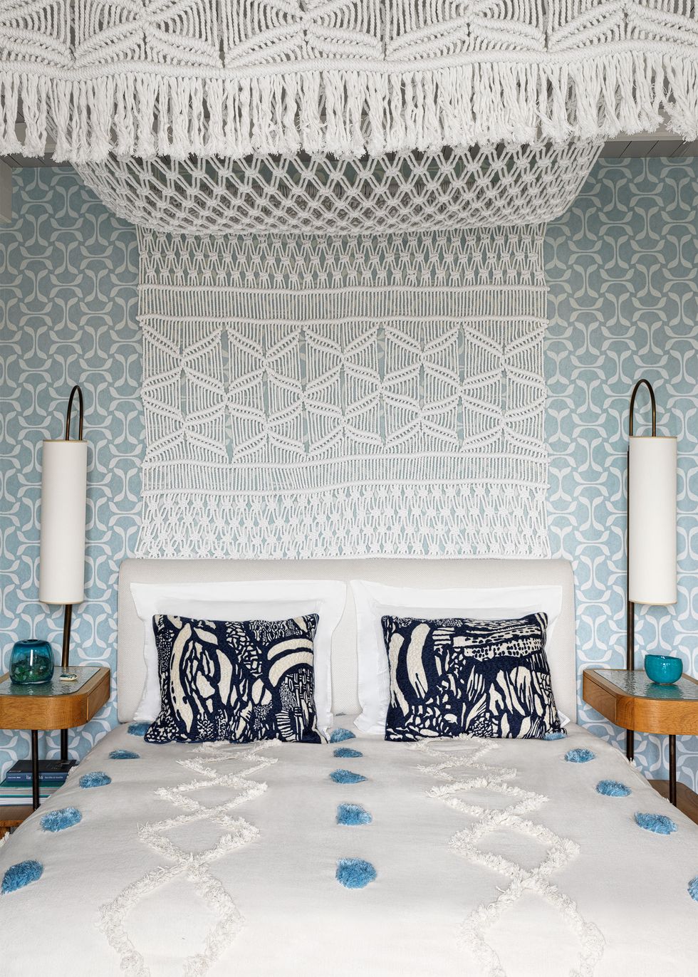 Bed with upholstered headboard and decorative pillows, white bedspread with blue pom-poms, nightstands with lamps on each side, crocheted bed canopy, wallpaper with a muted blue and white pattern