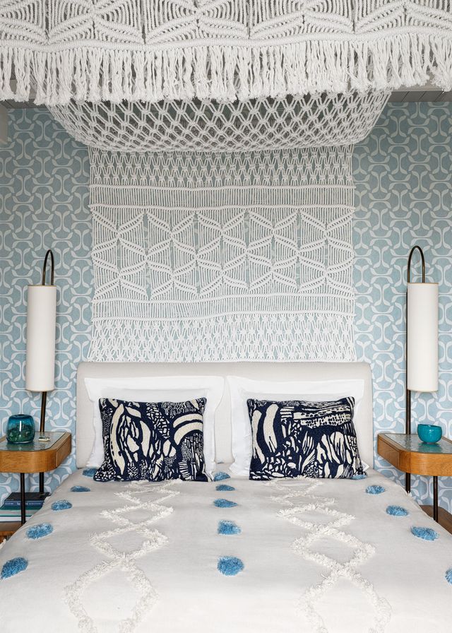 bed with upholstered headboard and decorative pillows, white bedspread with blue pompoms, nightstands with lamps on both sides, crocheted bed canopy, muted blue and white patterned wallpaper