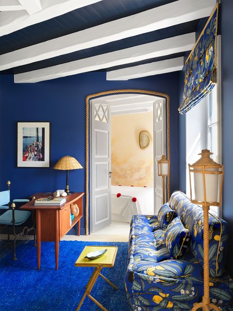 home office with a blue and yellow printed sofa and matching window shade, floor lamps, a wood scandinavian desk with an iron chair, bright blue area rug and deep blue painted walls, doors lead to a bedroom