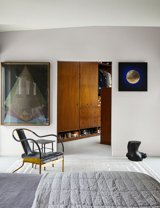 a bedroom has a bed with gray linen sheets and duvet, a geometric patterned area rug, a wood and leather armchair, a sculpture on the floor and two artworks on the wall by the door leading to the dressing room