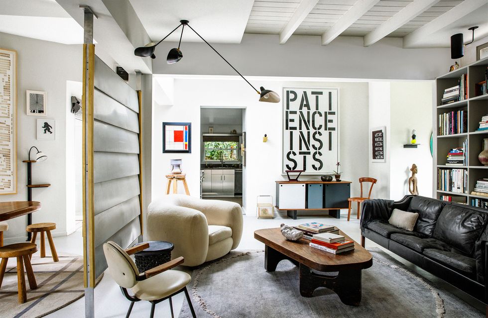 in the living room two light colored chairs face a black leather sofa with a wood cocktail table on an area rug, a console has an artwork that says patience insists, at left is a room divider and bookshelves line the right wall