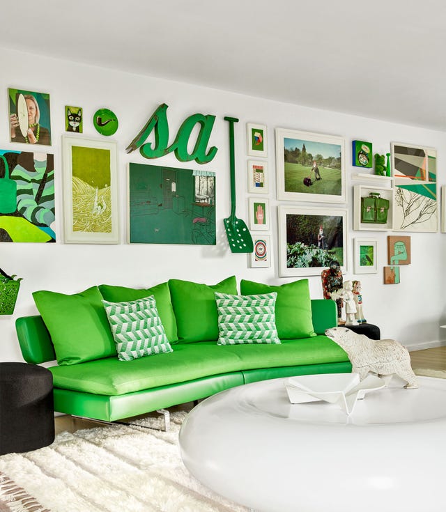 a studio room has an armless green sofa with patterned and solid green pillows, a ufo shaped cocktail table with a bear sculpture on it, numerous green themed artworks and objects adorn the wall
