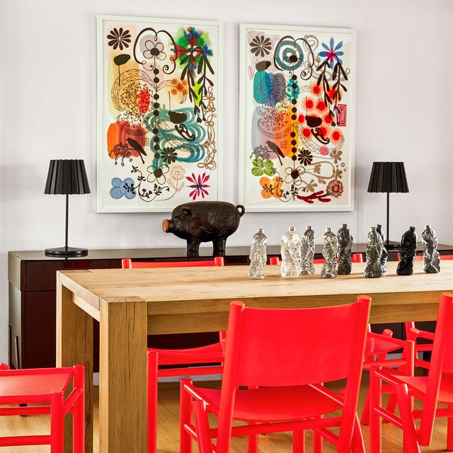 dining room has a wood table and on it are resin water bottles made to look like figures, red chairs, a console with two table lamps and a pig sculpture, two colorful abstract artworks hang above console