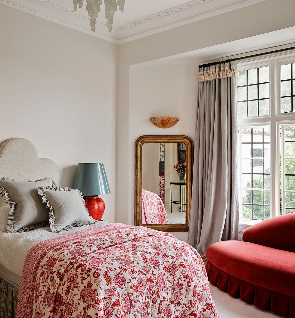in a child’s bedroom is a twin bed with a taupe fabric headboard and bedskirt and a printed red and white quilt, a lamp sits on a nightstand, a mirror is in the center and a red velvet daybed is in front of the window