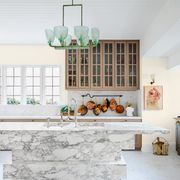 kitchen with marble island with sink, marble floor and counter and window and copper bottom pots hanging on the wall and glass paned cabinets above, a green tinted frosted glass pendant is above the island