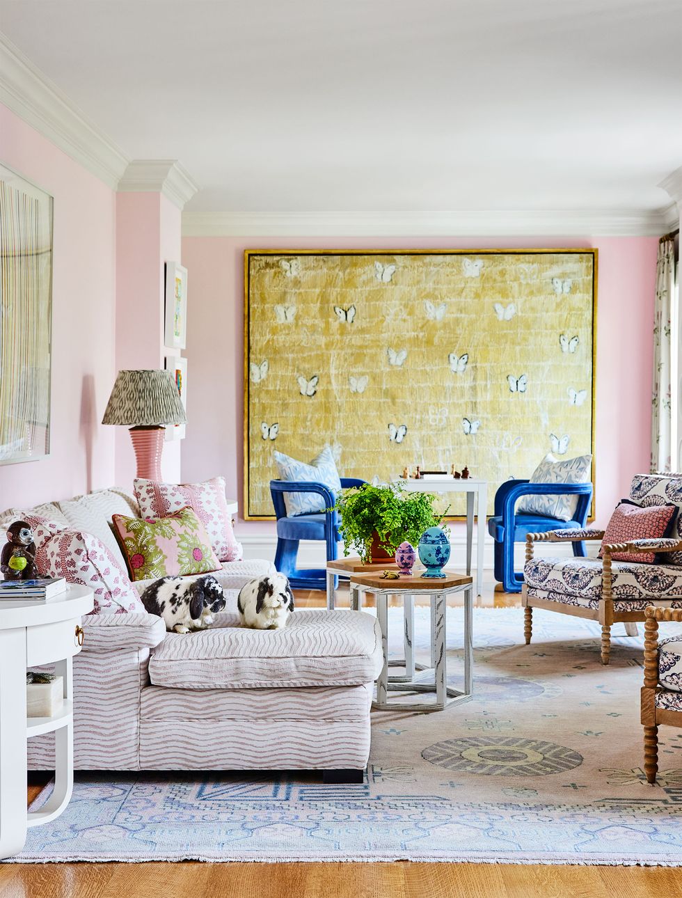 in the tv room with pink walls is an upholstered sofa, two chairs with carved wood arms and cocktail tables, and in the background are blue chairs and a large artwork with butterflies
