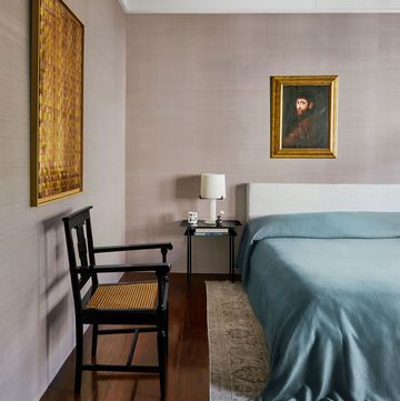 a bed with fabric headboard and a light blue linen cover, a black nightstand with a white lamp, a black lacquer wooden chair, a bleached kilim, and an old master painting and a woven metal artwork on walls